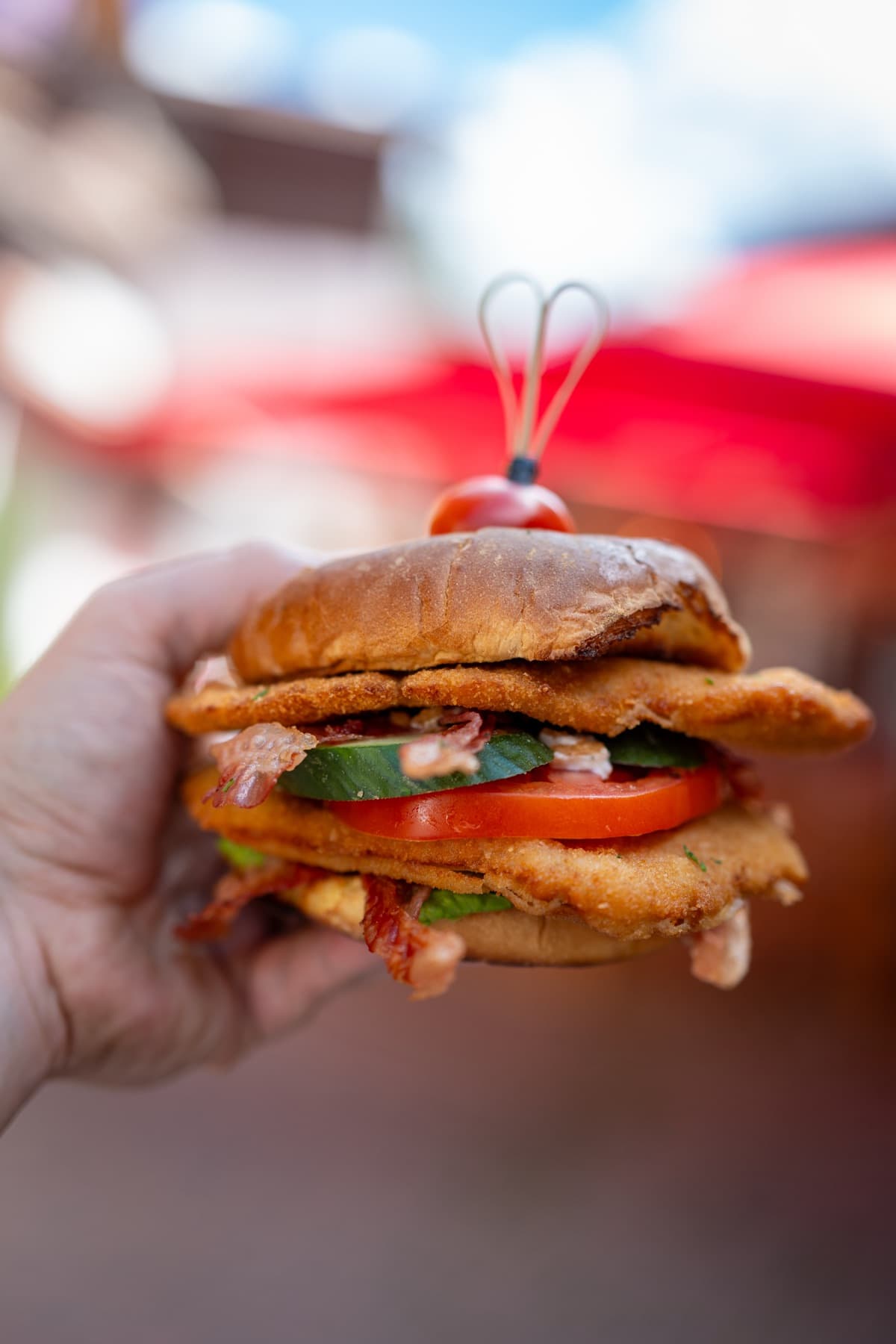 Schnitzel club burger with bacon, tomato and cucumber on a brioche bun, topped with a cherry tomato, held up in one hand