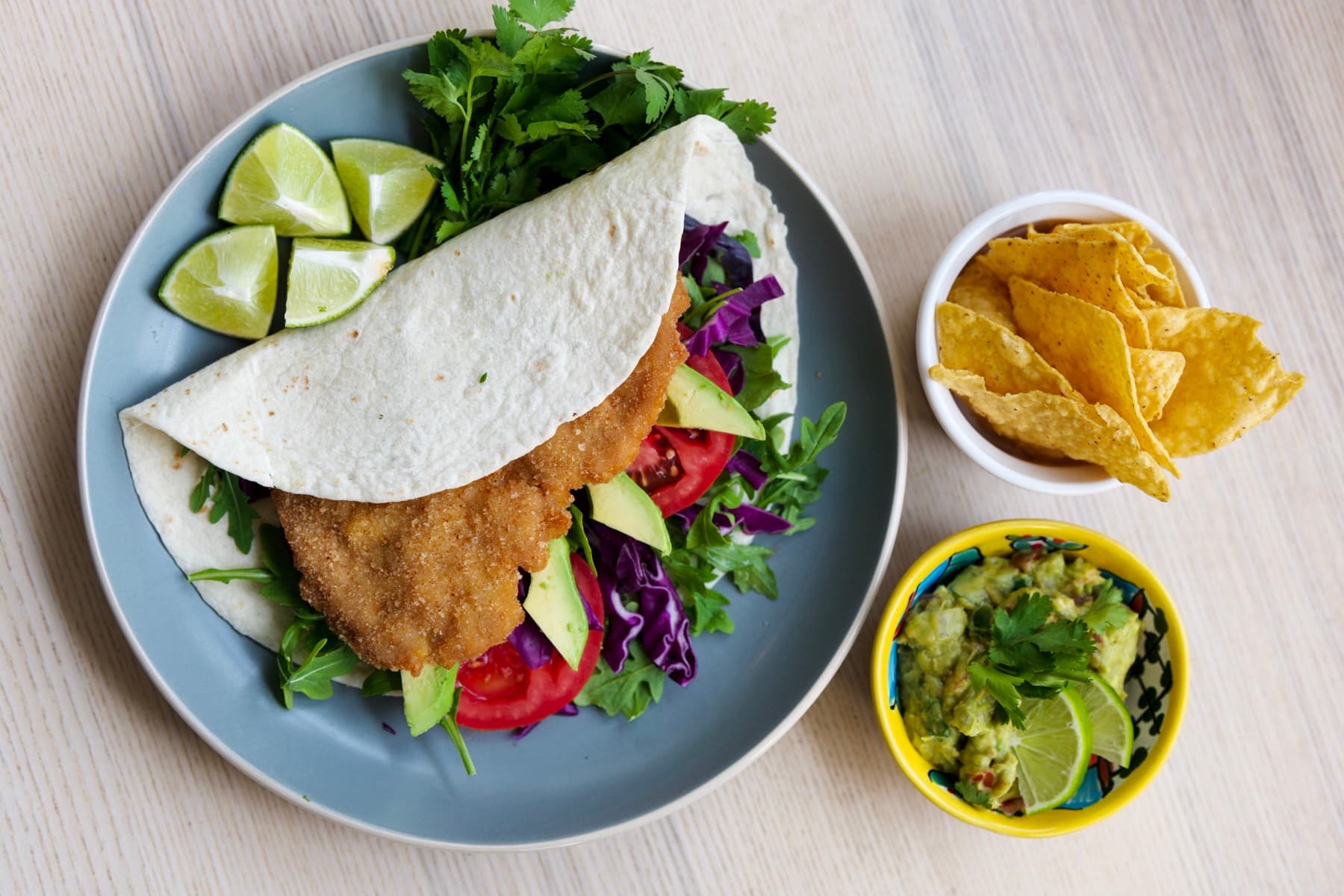 Schnitzel taco with avocado, tomato and arugula on a plate, served with tortilla chips and guacamole