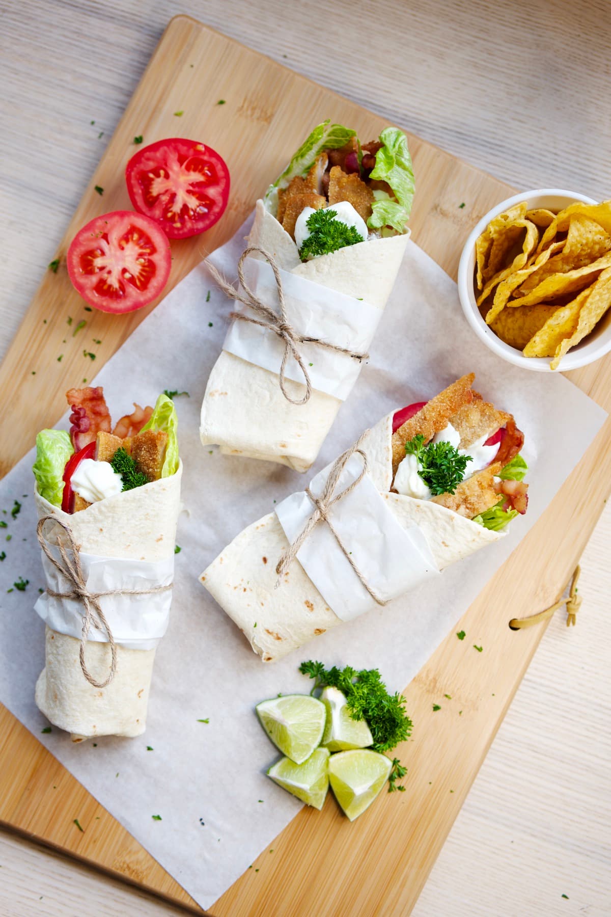 Three schnitzel wraps tied with twine on a wooden board, with tomatoes, lime and tortilla chips